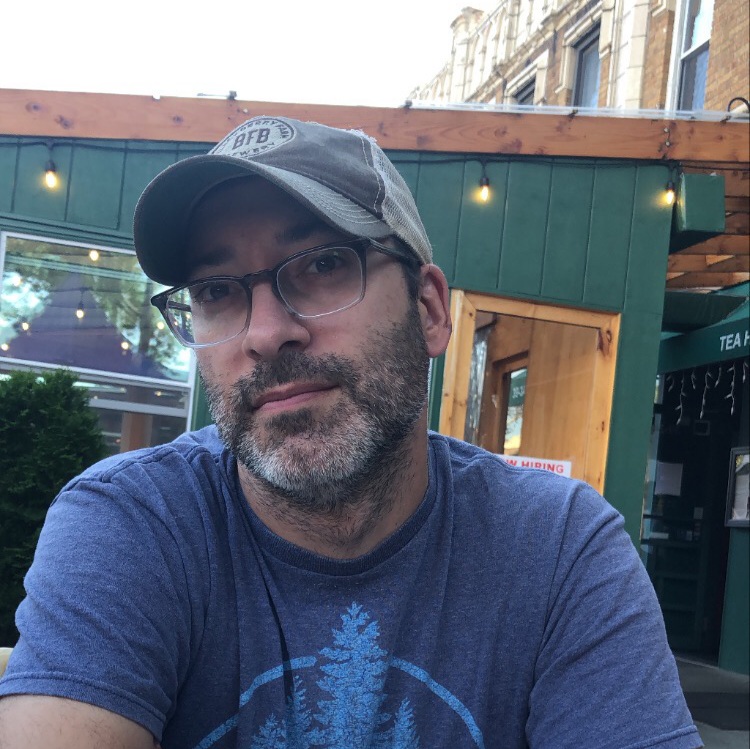 Photo of poet Jared Beloff. He's sitting outside. He has on a blue t-shirt and hat. With glasses and partly grey beard.