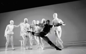 One dancer in black is surrounded by dancers in white