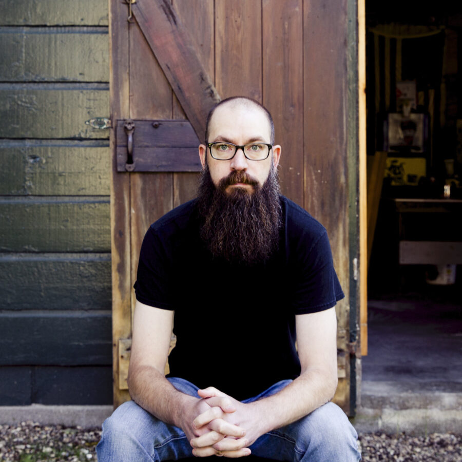 Author photo of poet Matthew Burns. He is sitting outside with his arms resting on his legs. He has on a dark black or blue t-shirt and blue jeans. He has glasses and a very long beard. Behind him is a door and a glimpse of something inside. 