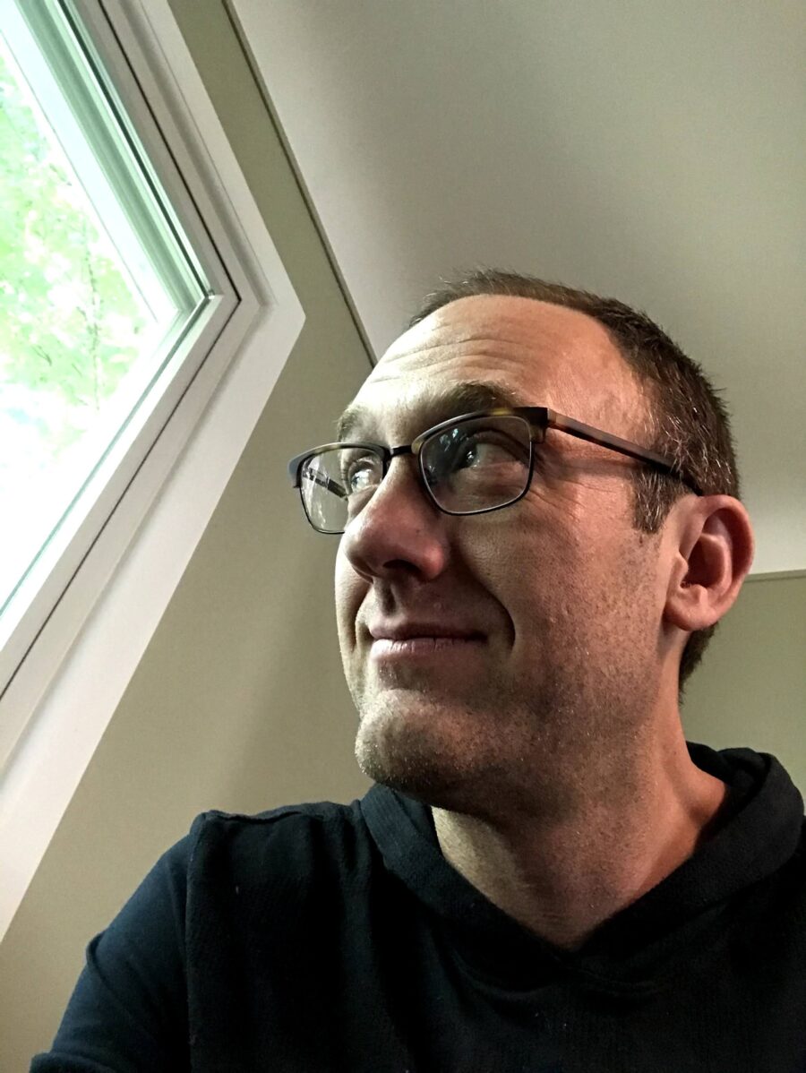 Color photo of poet and teacher Mitchell Nobis. He's inside a home, starring outside the window. He has glasses and a black shirt.