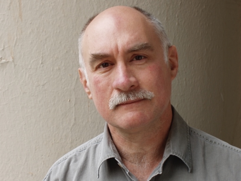 Headshot of poet and professor Al Maginnes. Al has white mustache and white hair on the side. He wears a gray shirt and is looking at the camera. Behind him is a grayish wall. 