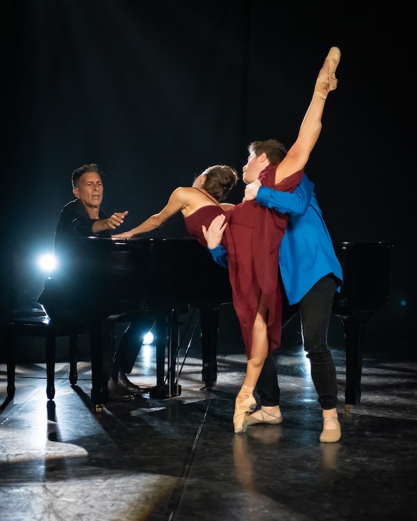 A dancer in red and a dancer in blue lean toward a pianist