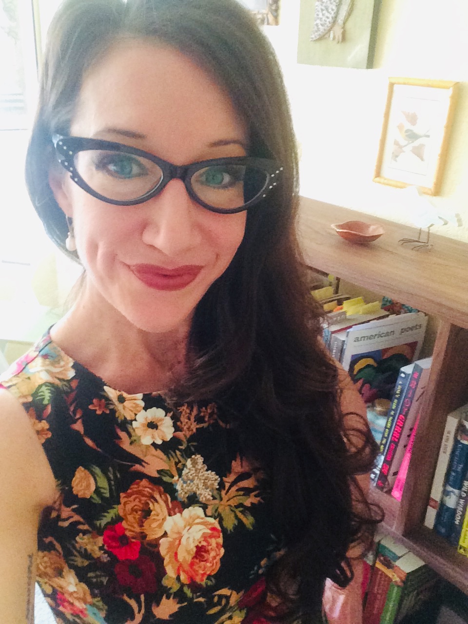Color photo of poet Rosemarie Dombrowski. She has fashionable eyeglasses. Her long dark hair is on her left shoulder. She's wearing a flower-print dress and there's a bookcase behind her. 