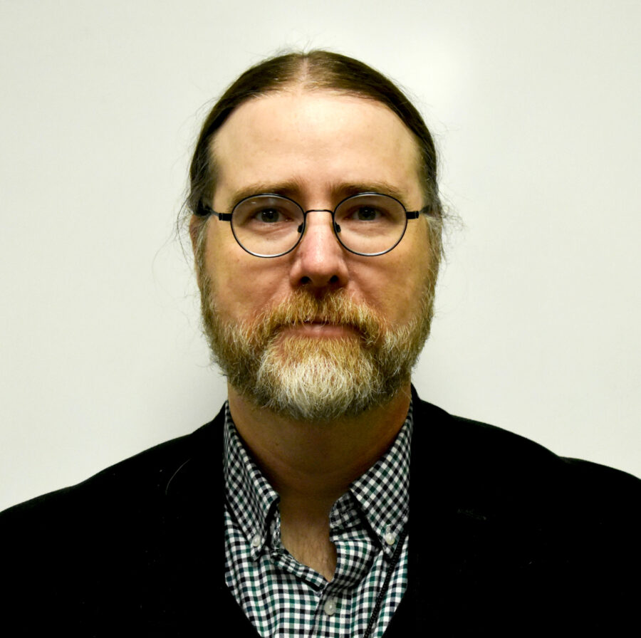 Headshot of poet and teacher Steve Henn. Steve has a trimmed beard; wears glasses; hair tied back. He has on a black jacket, with collar shirt underneath. 