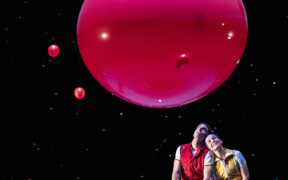 Two dancers stare at a large red balloon