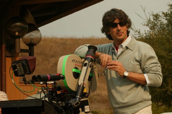 Alexander Payne standing, leaning on a film camera while wearing sunglasses outside.
