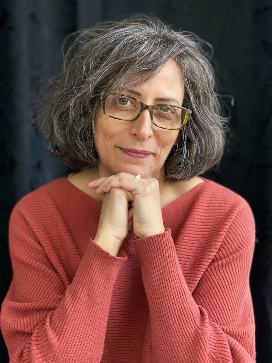 Color photo of poet and professor Julia Lisella. she's in a red sweater, her hands folded under her chin; she's wearing glasses and smiling with her lips closed.