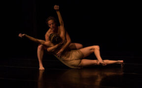 A dancer holds another dancer on floor with outstretched arms