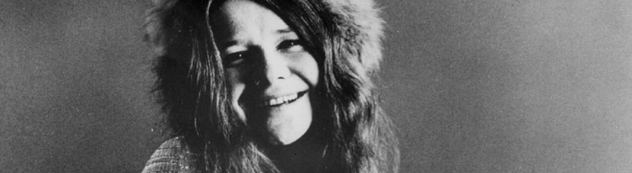 Janis Joplin smiling with a furry hat on in a black and white photo.