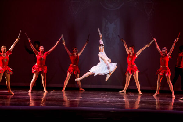 A dancer in white leaps before a group in red